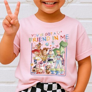 A young girl wearing a pink t-shirt with a colorful graphic of characters from the Toy Story movie, including Woody, Buzz Lightyear, and other beloved characters.