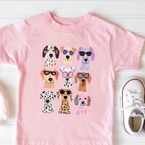 A pink t-shirt with a graphic of nine colorful dog faces wearing sunglasses, arranged in a 3x3 grid.