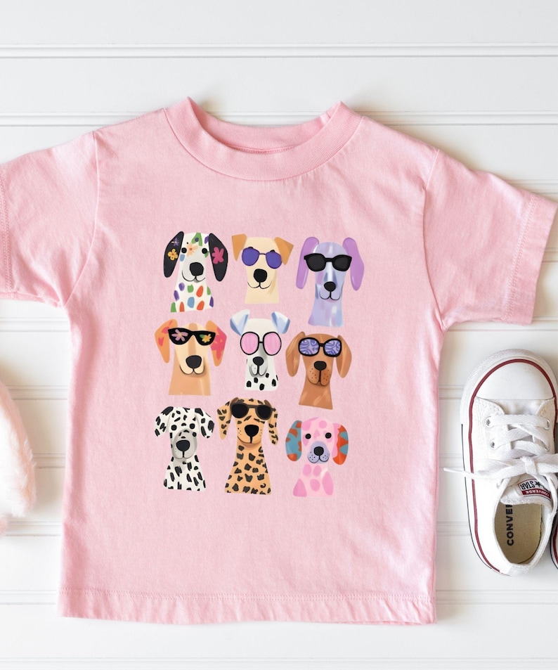 A pink t-shirt with a graphic of nine colorful dog faces wearing sunglasses, arranged in a 3x3 grid.