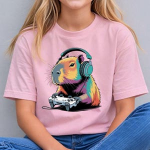 A woman wearing a pink t-shirt with a colorful cartoon image of a capybara wearing headphones and holding a video game controller.