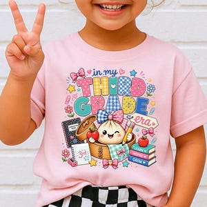 May include: A light pink t-shirt featuring a colorful graphic with the text "in my THIRD GRADE era". The design includes a smiling dumpling in a bamboo steamer, a composition book, a notebook labeled "3rd", and a stack of books labeled "LEARN INSPIRE GROW".