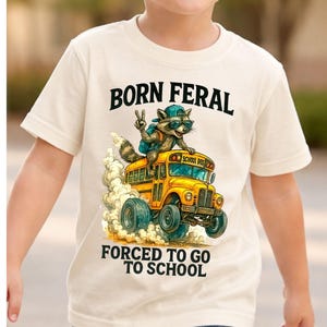 A young boy wearing a white t-shirt with a graphic design of a raccoon riding a school bus and the text "Born Feral Forced to go to school".