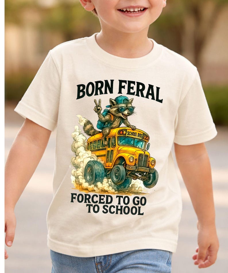 A young boy wearing a white t-shirt with a graphic design of a raccoon riding a school bus and the text "Born Feral Forced to go to school".