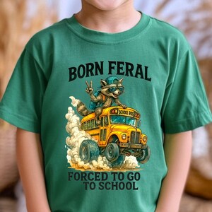A young boy wearing a green t-shirt with a graphic design of a raccoon riding on the back of a school bus, along with the text "Born Feral Forced to Go to School".