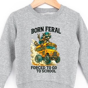 A gray sweatshirt with a graphic design depicting a raccoon riding on the back of a school bus, with the text "BORN FERAL FORCED TO GO TO SCHOOL" printed on it.