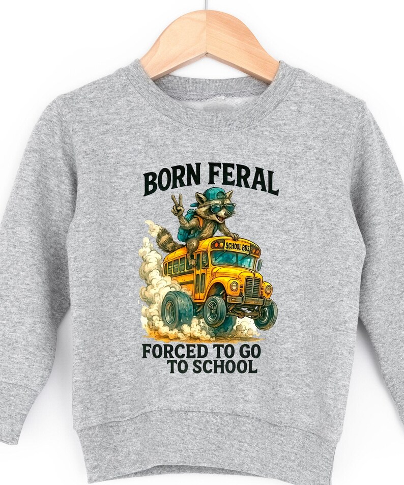 A gray sweatshirt with a graphic design depicting a raccoon riding on the back of a school bus, with the text "BORN FERAL FORCED TO GO TO SCHOOL" printed on it.