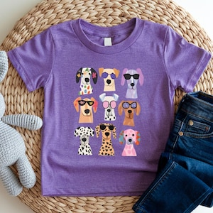 A purple t-shirt with a graphic of nine colorful dog faces wearing sunglasses, arranged in a 3x3 grid. The t-shirt is placed on a woven placemat, and there is a pair of blue jeans nearby.