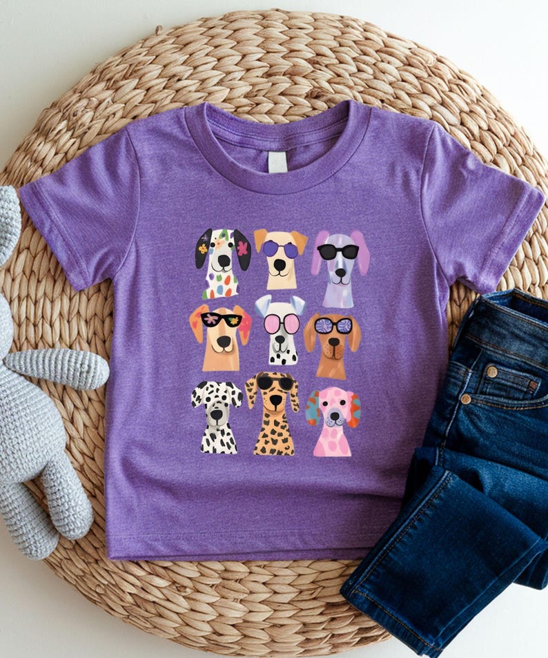 A purple t-shirt with a graphic of nine colorful dog faces wearing sunglasses, arranged in a 3x3 grid. The t-shirt is placed on a woven placemat, and there is a pair of blue jeans nearby.