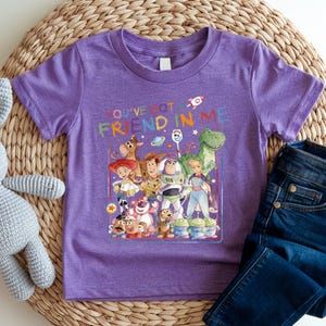 A purple t-shirt with a colorful graphic of characters from the Toy Story movie, along with the text "You've got friend in me" printed on it. The t-shirt is placed on a woven mat, and there is a pair of jeans visible in the background.