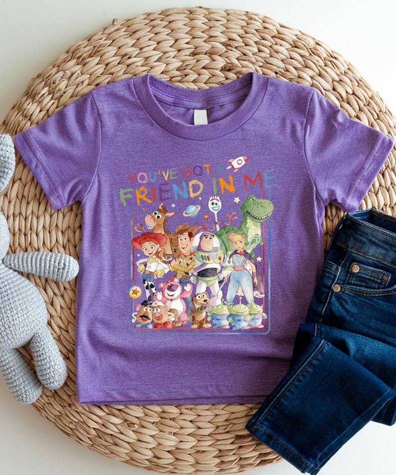 A purple t-shirt with a colorful graphic of characters from the Toy Story movie, along with the text "You've got friend in me" printed on it. The t-shirt is placed on a woven mat, and there is a pair of jeans visible in the background.