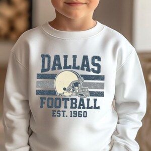 A young boy wearing a white sweatshirt with a graphic design featuring the Dallas Cowboys football team logo and text.