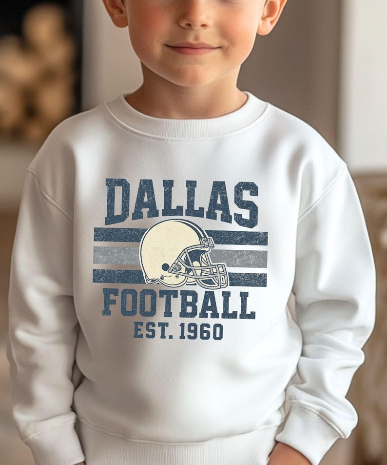 A young boy wearing a white sweatshirt with a graphic design featuring the Dallas Cowboys football team logo and text.