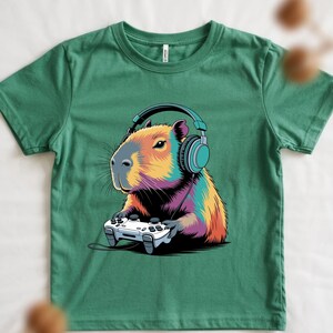 A green t-shirt with a colorful illustration of a capybara wearing headphones and holding a video game controller.