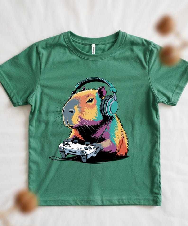 A green t-shirt with a colorful illustration of a capybara wearing headphones and holding a video game controller.