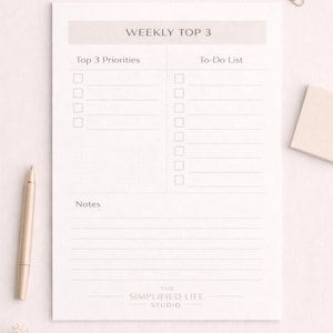 May include: A weekly planner notepad with the text "WEEKLY TOP 3" at the top. It has sections for "Top 3 Priorities" and "To-Do List", with space for notes. A gold pen, paper clips, and a small notepad are also present.