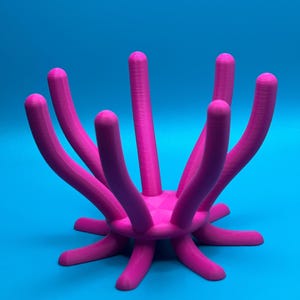 May include: A vibrant magenta-colored, abstract sculpture with a central geometric base and multiple curved, cylindrical arms extending outwards. The sculpture is set against a bright blue background, creating a striking contrast. The design is modern and artistic.