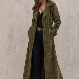 May include: An olive green suede duster coat with a notched collar and button-front closure, worn over a black V-neck top and black jeans with a brown leather belt featuring a large metallic buckle, paired with brown leather western-style boots.