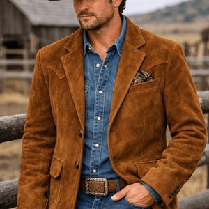 Könnte beinhalten: Ein schwarzer Cowboyhut mit silbernem Nietband, ein brauner Wildlederblazer mit steigendem Revers und Pattentaschen, ein blaues Jeanshemd sowie ein brauner Ledergürtel mit einer großen, verzierten, rechteckigen silberfarbenen Metallschnalle.