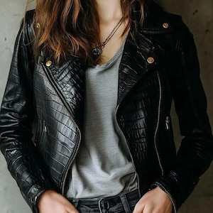 Könnte beinhalten: Schwarze Lederjacke mit einem strukturierten, krokodilartigen Muster. Die Jacke hat einen diagonalen Reißverschluss, goldfarbene Knöpfe und einen klassischen Biker-Stil. Das Model trägt ein graues T-Shirt und dunkle Jeans.