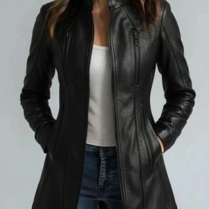 May include: A black leather jacket with a stand-up collar and multiple zippered pockets. The jacket has a tailored fit and a long length, reaching mid-thigh. The model is wearing a white top and blue jeans.