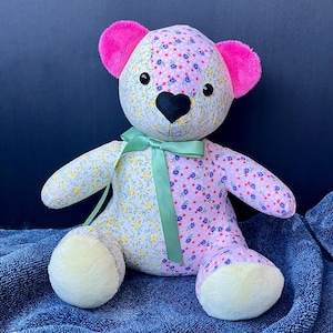 May include: A handmade teddy bear with a floral pattern in pink, yellow, and blue. The bear has pink ears, a black heart-shaped nose, and a green ribbon. The bear is sitting on a blue blanket.