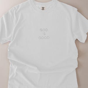 May include: White t-shirt with the text "GOD IS GOOD" in the center. The shirt is made of a soft material and has a crew neck. The word "WHITE" is in the bottom right corner.