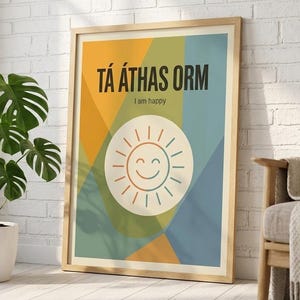 May include: A framed art print with the text "TÁ ÁTHAS ORM" and "I am happy". The artwork features a smiling sun graphic in a circle, set against a geometric background of orange, green, and blue. The frame is a light wood color.