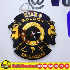 May include: A black and gold wall clock with the words "RIAN G SALOON" and "SHAHZAB" in gold lettering. The clock features two gold-colored barber profiles and a clock face with gold numerals. Barber tools are also displayed.