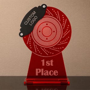 Op de afbeelding: Een rode acryltrofee in de vorm van een remschijf met een zwarte remklauw aan de bovenkant. De remklauw is voorzien van de tekst CUSTOM LOGO en de voet van de trofee is gegraveerd met de tekst 1st Place in een duidelijk schreefloos lettertype.