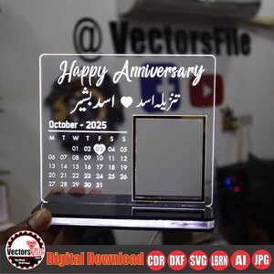 May include: A clear acrylic anniversary plaque with the words "Happy Anniversary" and text in a foreign language. The plaque includes a calendar for October 2025 and a photo frame. The base has a black finish with a glowing light.