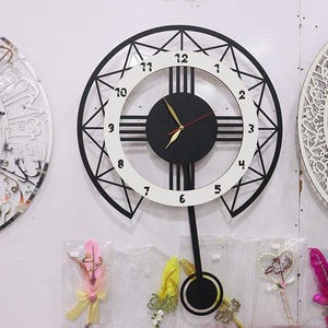 Puede incluir: Un reloj de pared en blanco y negro con péndulo, con un diseño circular con números romanos. El reloj tiene una esfera y manecillas negras, y está rodeado por un patrón geométrico. Se ven otros objetos decorativos.