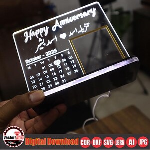 May include: A black acrylic anniversary calendar with the words "Happy Anniversary" and a calendar for October 2025. The calendar has a built-in light and a small rectangular frame. The text is in white.