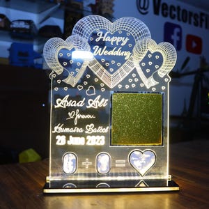 May include: A clear acrylic wedding decoration with a heart-shaped design. The text "Happy Wedding" is at the top, with names and a date below. A glittery green square is in the center, and a heart-shaped design is at the bottom.