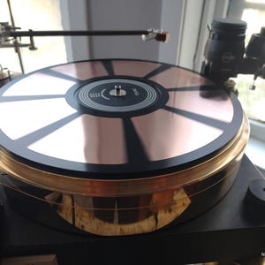 Könnte beinhalten: Hochwertiger Plattenspieler mit kupferfarbenem Plattenteller und schwarzem Sockel. Der Plattenteller hat ein radiales Design mit schwarzen Akzenten. Der Plattenspieler verfügt über goldfarbene Zierleisten und einen Tonarm. Das Design ist elegant und modern.