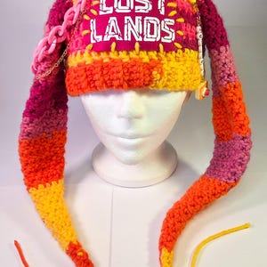 May include: A multi-coloured crochet hat featuring long ear flaps, a pink rectangular patch with the white text LOST LANDS, a chunky pink plastic chain, and a dangling beaded charm. The hat is crafted in shades of orange, yellow, and magenta.