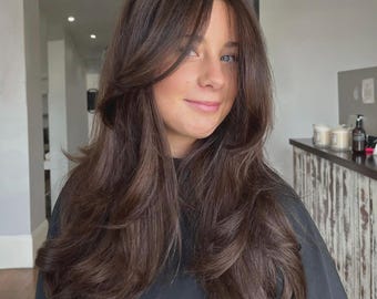 Può includere: Una persona con capelli lunghi castano scuro acconciati con strati voluminosi e una frangia a tendina con riga centrale. I capelli hanno una consistenza liscia e lucida con onde morbide sulle punte, che incorniciano il viso e scendono verso il basso.