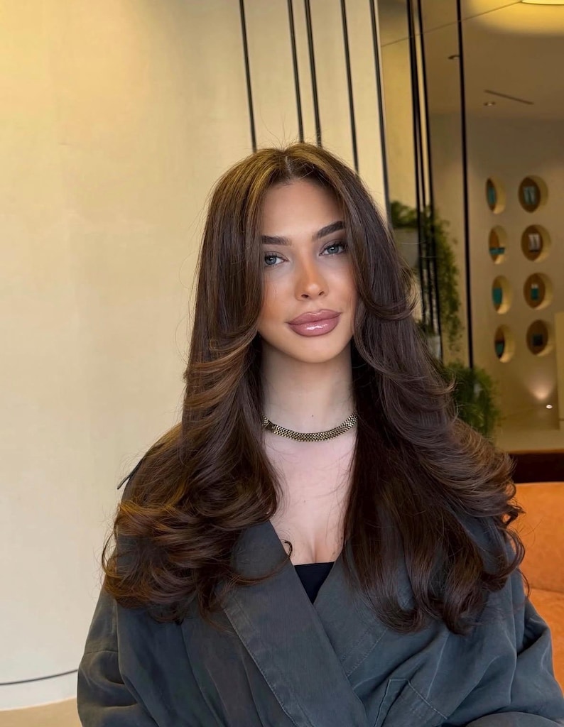 May include: A person with long, dark brown hair styled in voluminous layers with a center part and face-framing curtain bangs. The hair features subtle lighter brown highlights throughout. A gold-toned, textured chain necklace is worn around the neck.
