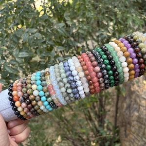 May include: A collection of beaded bracelets in various colors, including black, orange, turquoise, white, and green. The bracelets are displayed on a gray cylindrical holder with a gold rim, showcasing a variety of natural stone beads.