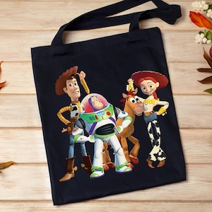 May include: A navy blue tote bag featuring a colorful graphic of Woody, Buzz Lightyear, and Jessie from Toy Story. The bag has black handles and is made of a durable fabric, perfect for carrying books or groceries.