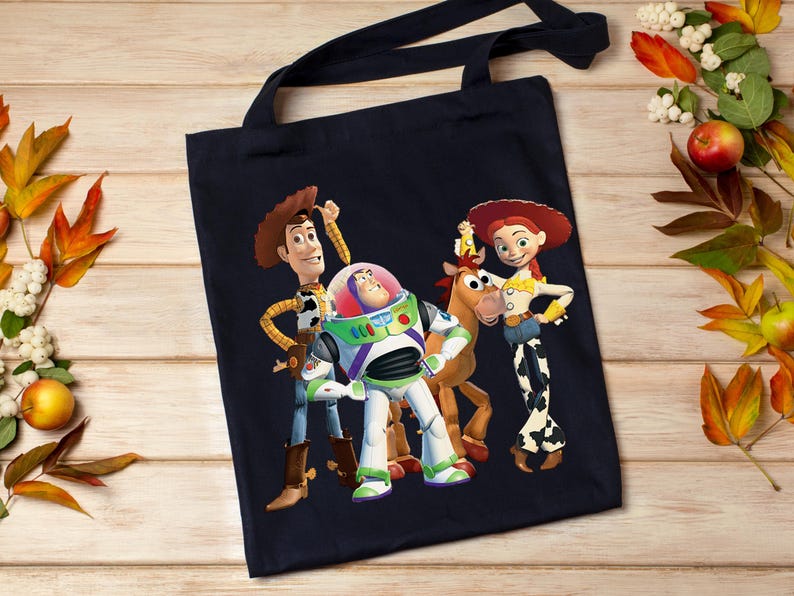 May include: A navy blue tote bag featuring a colorful graphic of Woody, Buzz Lightyear, and Jessie from Toy Story. The bag has black handles and is made of a durable fabric, perfect for carrying books or groceries.