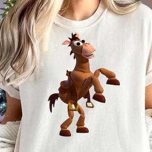 May include: A white t-shirt featuring a cartoon image of a brown horse with a saddle. The horse is in a standing position with its front legs raised. The shirt has short sleeves and a crew neck.