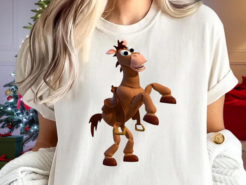 May include: A white t-shirt featuring a cartoon image of a brown horse with a saddle. The horse is in a standing position with its front legs raised. The shirt has short sleeves and a crew neck.
