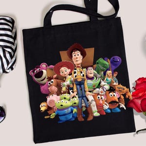 May include: Black tote bag featuring a colorful graphic of characters from the animated film Toy Story. The bag has black handles and is made of a sturdy material. A cup of coffee, sunglasses, and a bouquet of red roses are also in the image.