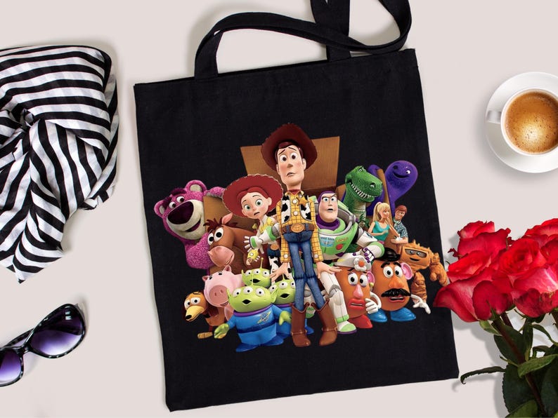 May include: Black tote bag featuring a colorful graphic of characters from the animated film Toy Story. The bag has black handles and is made of a sturdy material. A cup of coffee, sunglasses, and a bouquet of red roses are also in the image.