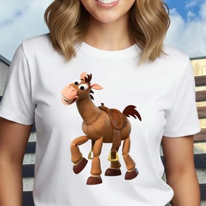 May include: White t-shirt featuring a cartoon image of a brown horse with a saddle. The horse has a friendly expression and is in a standing position. The shirt is made of a soft material and has short sleeves. The background is a blurred outdoor scene.