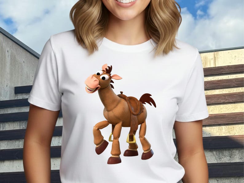 May include: White t-shirt featuring a cartoon image of a brown horse with a saddle. The horse has a friendly expression and is in a standing position. The shirt is made of a soft material and has short sleeves. The background is a blurred outdoor scene.