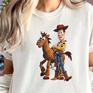 May include: White t-shirt featuring a cartoon image of Woody from Toy Story standing next to a brown horse. Woody is wearing a cowboy hat, yellow plaid shirt, blue jeans, and brown boots. The horse has a brown mane and tail.