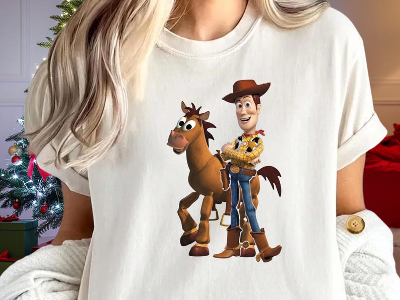 May include: White t-shirt featuring a cartoon image of Woody from Toy Story standing next to a brown horse. Woody is wearing a cowboy hat, yellow plaid shirt, blue jeans, and brown boots. The horse has a brown mane and tail.
