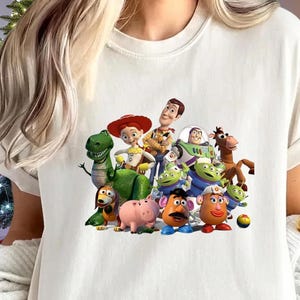 May include: Cream-colored t-shirt featuring a colorful graphic of characters from the animated movie "Toy Story." The design includes Woody, Buzz Lightyear, and other beloved characters. The shirt has short sleeves and a crew neck.