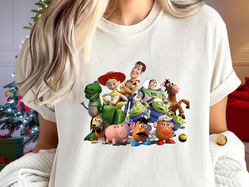 May include: Cream-colored t-shirt featuring a colorful graphic of characters from the animated movie "Toy Story." The design includes Woody, Buzz Lightyear, and other beloved characters. The shirt has short sleeves and a crew neck.